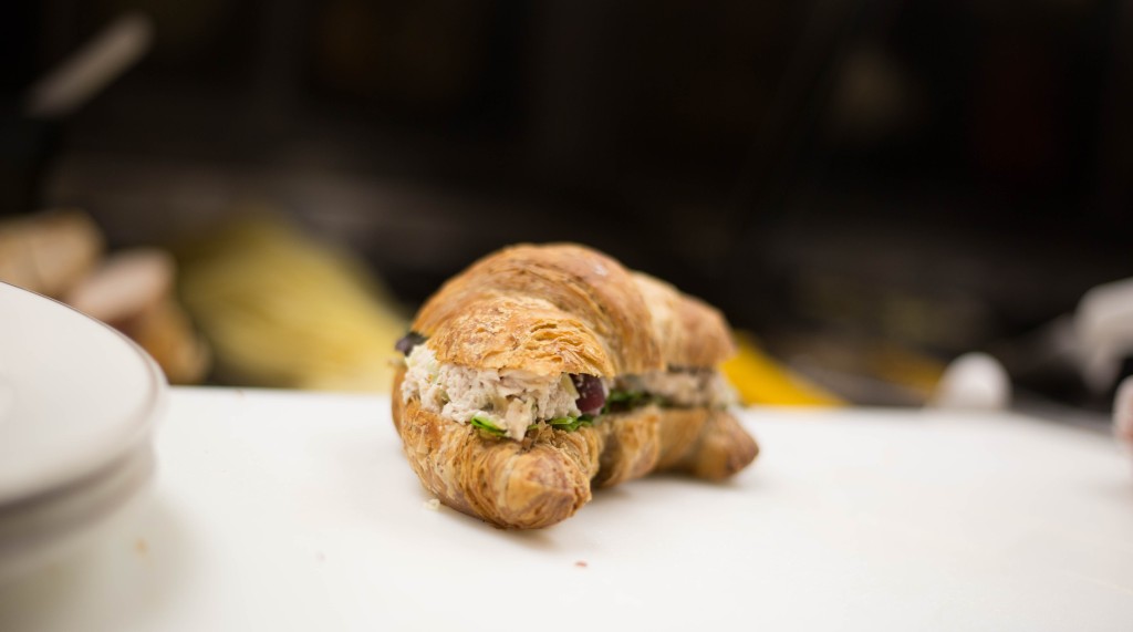 Turkey salad croissant sandwich sits alone on a counter top beside a short stack of plates. Behind, a dark room. 